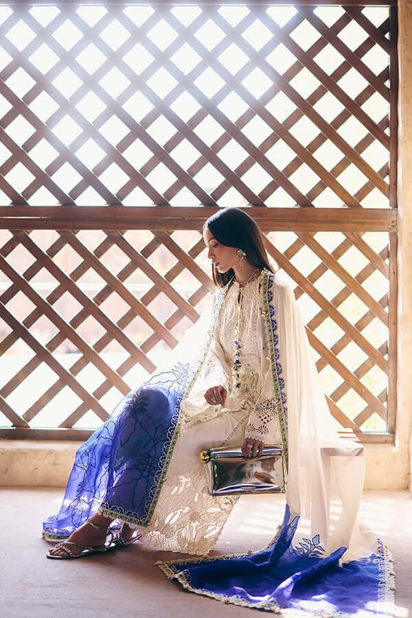 Model wearing Zehra outfit with ivory embroidery and blue floral patterns, seated near a lattice window.